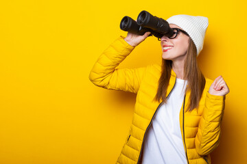 Young woman in a yellow jacket smiling looks through binoculars on a yellow background.