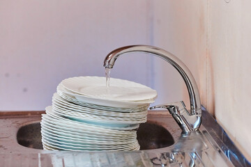 A pile of dirty dishes stands under a running water tap in a steel sink