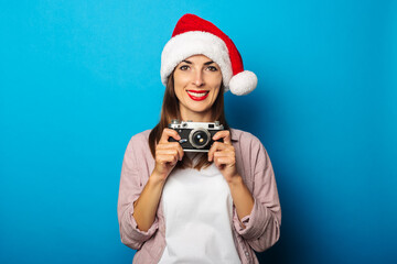 Smiling young woman in a shirt wearing a Santa Claus hat holding a retro camera on a blue background.