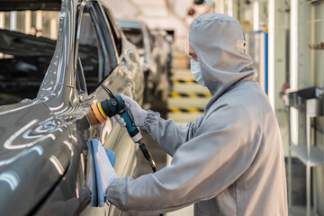 An employee of the car body paint shop with a medical mask on his face polishes the painted surface