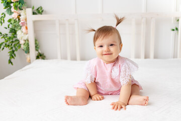 cheerful baby girl six months old sitting in a bright beautiful room on a white bed in pink clothes and smiling