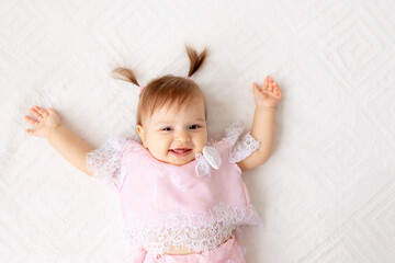 portrait of a little baby girl six months old on a white bed in pink clothes, holding her hands up and smiling
