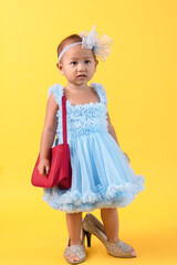 Lovely Asian infant girl dressing dress with red bag and wearing adult high heel shoes and ties up bow headband on yellow background