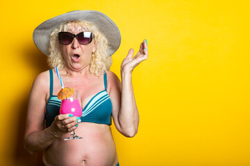 Old woman in a swimsuit with a hat holding a cocktail in surprise on a yellow background.