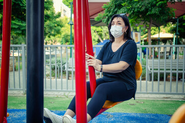 Mature woman in a face mask attends an urban outdoor gym in a park near her during the pandemic