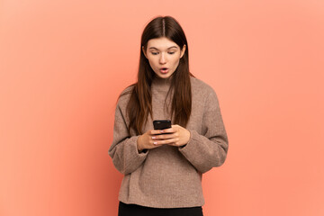 Young Ukrainian girl isolated on pink background looking at the camera while using the mobile with surprised expression