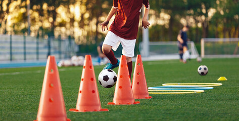 Naklejka premium Boys running soccer balls between training cones. European football sports summer camp for youth athletes. Soccer boys with balls on grass pitch. Soccer goal and stadium in the background