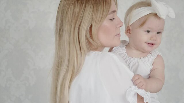 Portrait of mom and cute little girl. A baby with blue eyes, in delicate clothes with a bow on her hair, looks at the camera. A young mother holds her baby on the arms