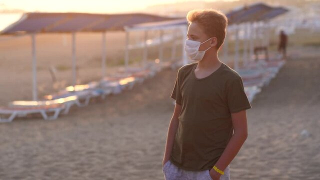 Young white kid standing on beautiful sunny sunset beach alone wearing medical mask on his face because of covid 19 lockdown
