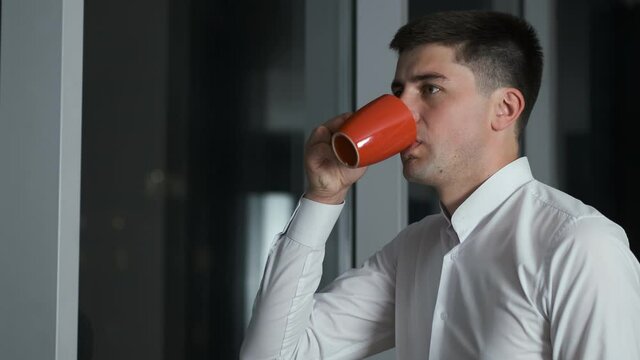 A 30-year-old businessman in a shirt stands in his office near a panoramic window overlooking the night city. He thinks about a business plan and drinks coffee and tea