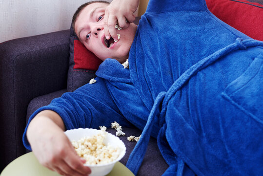 Fat Man In A Robe Eating Popcorn While Lying On The Couch.