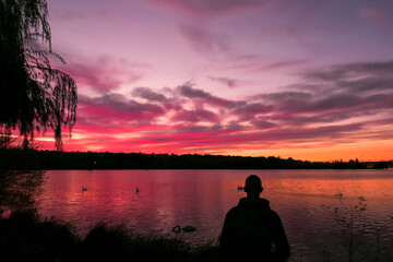 Fototapeta premium Silhouette of a man in front of the water with amazing sunset. Beautiful landscape with a lake and dramatic sky with cumulus clouds on the horizon. Weeping willow in the foreground.