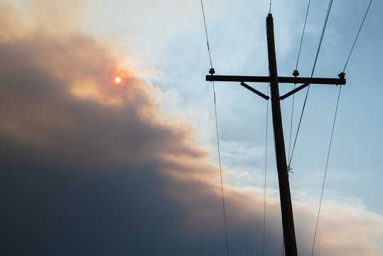 Smoke plume from the Cameron Peak fire in Colorado  fills the sky and partially obscures the sun. A wooden pole holding electric wires is in the foreground. 