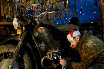 Fototapeta na wymiar A man in a santa claus hat repairs a motorcycle in the garage on New Year's Eve.