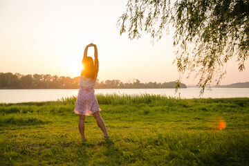 A beautiful girl raised her hands on the bank of the river, in the rays of the setting sun. Fresh air, unity with nature.