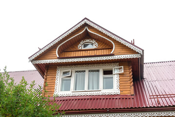 Obraz premium Retro mansard with large decorated window on red roof of traditional Russian country house in small village against cloudy sky on nice sunny day low angle shot.