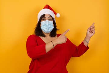Fototapeta na wymiar Beautiful middle aged woman wearing Christmas hat and medical mask standing against yellow background, pointing aside at copy space. Protection against infectious disease.
