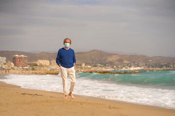 Senior pensioner relaxed in beach during new normal - retired old man on his 70s wearing surgical mask against covid19 walking at the sea thoughtful and contemplative