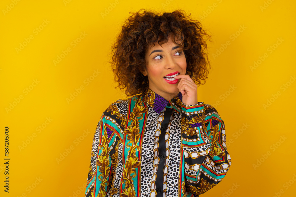 Sticker Dreamy female with thoughtful expression, looks to the camera, keeps hand near face, bitting a finger thinks about something pleasant, poses against gray wall.