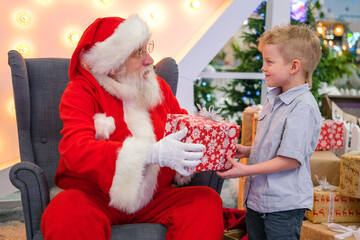 Fototapeta na wymiar Santa Claus gives boy a gift in Shopping Mall. Real authentic Santa talking and playing surprise games with kids . Christmas sales and wishes.