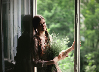 Young woman with two braids in simple dress holding flowers near window