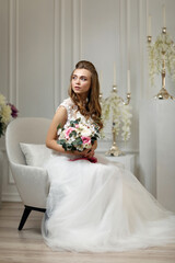 Gorgeous tender young bride with flower bouquet sitting on armchair in white studio interior