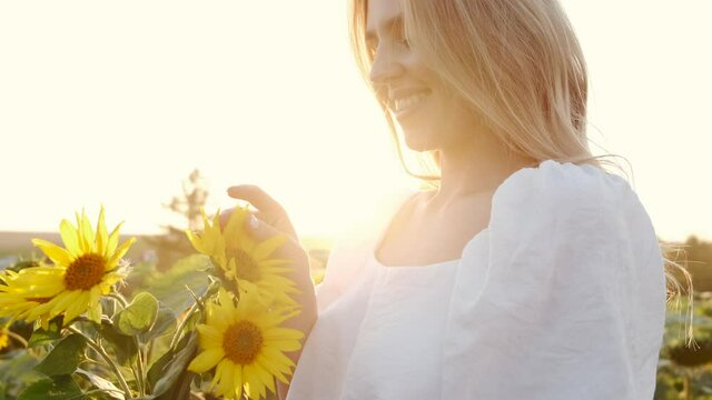 Beautiful young woman standing in field with sunflowers and enjoying nature. Woman smells flowers against background of nature and field of sunflowers. Portrait view. 4k.