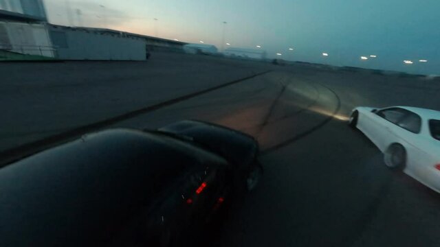 Sports automobiles drive fast drifting smoke from wheels on empty plain road in contemporary night city first point view fpv sport drone follow close shot, round evening sunset panorama