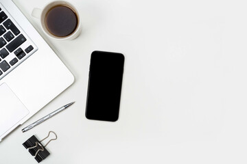 a phone with a screen mockup, a coffee cup of writing supplies, a pens, a notepad on a white wooden table background. Top view of the work area, copy space