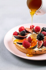 Bagel with cream cheese, figs, raspberries, blackberries and honey.  Close-up of gourmet sandwich on pink plate on gray background.