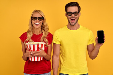 Excited couple friends guy and girl in 3d glasses isolated on yellow background. People in cinema concept. Watching movie film, hold bucket of popcorn, mobile phone with blank empty screen