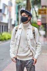 Stylish man wearing mask and walking down the street wearing white sweater and jacket standing on street