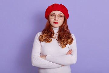 Portrait of lovely woman wearing white shirt and red beret, looking at camera with serious facial expression, stands with folded hands, looking strict at somebody.