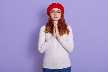 Close-up portrait of nice attractive pretty girl keeping arms together asking favor, expecting luck isolated over lilac pastel background, female wearing red beret, winsome woman with wavy hair.