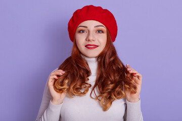 Young beautiful girl with red lips stands in red beret and white shirt, posing isolated over lilac background, looks at camera and smiling, touching her curls with palms.