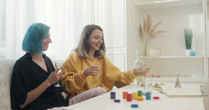 Two young women in a same-sex lesbian relationship sitting down to enjoy a glass of white wine together at home at a table with colorful pots of paint to draw the LGBT rainbow symbol
