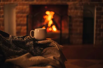 Fototapete Tee Mug with hot tea standing on a chair with woolen blanket in a cozy living room with fireplace.  © polinaloves