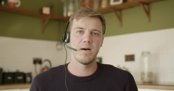 Male teacher using headset during video call and looking at camera, working from home at kitchen table during pandemic