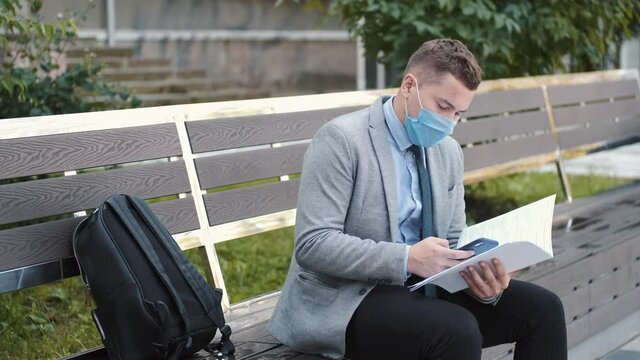 Freelancer. The man in disposable mask read the papers sitting on a bench in a city park. Coronavirus. Social distane.