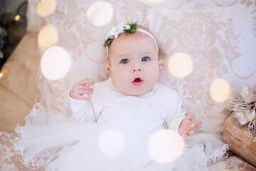 Fototapeta premium Close-up portrait of a little baby in a white dress with an ornament on her head. The girl lies on a light sofa in garlands of lights, a festive Christmas tree, laughing. Copy space, greeting card