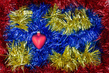 Christmas toy in the form of heart on a background of blue, red and yellow tinsel. It looks like a...