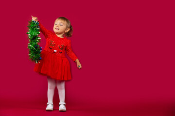 Little happy girl in a red New Year's dress, with a green fluffy garland in her hands. Holiday concept on red background