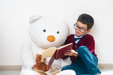 A schoolboy in a sweater and glasses shows an open book to a large teddy bear. Conceptual. Copy space.