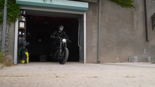 Slowmotion video of motorcyclist in a black helmet rides a classic motorcycle out of a garage near his house. Footage. The bike drove from a workshop on the first floor of the house.