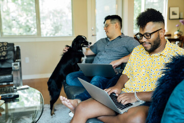 Happy gay couple working from home with laptops on couch with their Australian Shepherd 