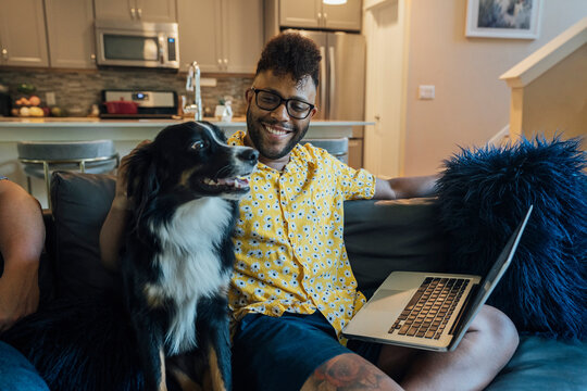 Man working from home with laptop sitting on couch and petting happy Australian Shepherd pet dog