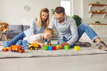 Concerned little boy makes colored cubes with his parents sitting on the carpet in the room.