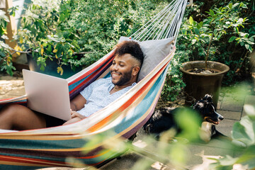 Man relaxing in hammock working on laptop computer  - obrazy, fototapety, plakaty