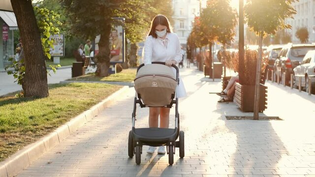 Caring mother in a protective mask is wakking with a baby in a pram during pandemic. The woman is taking care of a child and giving her love.