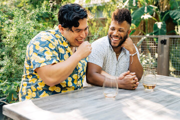 Happy gay couple sitting in back yard drinking wine together and laughing. 